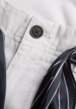 Gros plan sur la ceinture d'un pantalon blanc avec un bouton noir portant l'inscription "Superdry" et un tissu bleu marine adjacent avec des rayures roses et blanches.