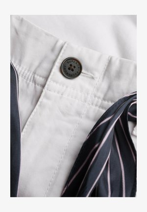 Zbliżenie na pas białych spodni z czarnym guzikiem oznaczonym napisem "Superdry" oraz przylegającą granatową tkaniną z różowo-białymi paskami.