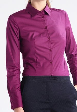 Femme portant une chemise magenta ajustée à manches longues, boutonnée et rentrée dans un pantalon sombre à motifs, debout avec une main dans la poche.
