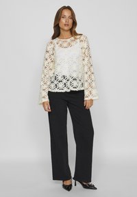 Haut en dentelle avec motif floral, couleur blanche, tissu transparent, manches longues. Associé à un pantalon noir texturé et des escarpins noirs à bout pointu.