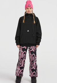 Zwarte pullover met een voorzak en verstelbare nek, gecombineerd met zwarte broeken met een abstract patroon in roze en zilver. Roze geribde beanie.