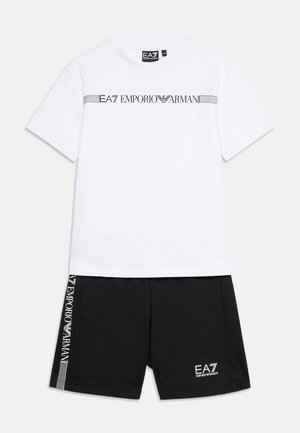 Biała koszula z krótkim rękawem EA7 Emporio Armani z czarnym logo na klatce piersiowej, zestawiona z czarnymi szortami z pionowymi i dolnymi białymi logo.