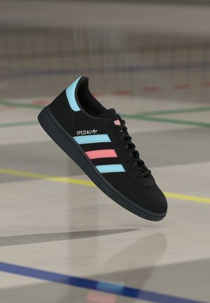 Svart Adidas-sneaker med blå og rosa striper og teksten "SPEZIAL", svevende over et gymgulv med fargede baner.