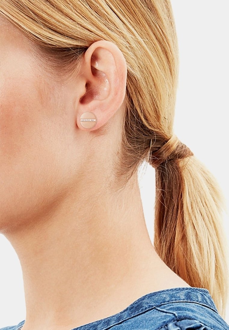 Femme blonde aux cheveux attachés portant des clous d'oreilles ronds beiges avec une bande horizontale de petits cristaux.