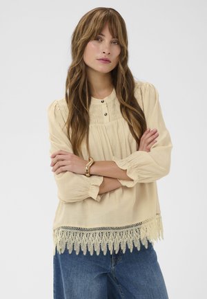 Kobieta o długich brązowych włosach, ubrana w kremową bluzkę z koronkowym dołem, niebieskie dżinsy i złotą bransoletkę, stoi z założonymi ramionami.