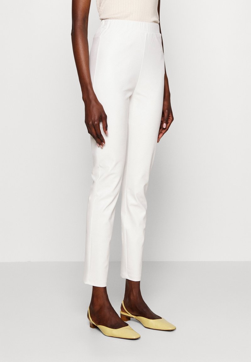 Femme portant un pantalon slim blanc taille haute et des chaussures slingback à talon bas jaune pastel, debout sur un fond uni.