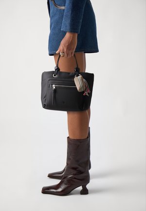 Personne portant des bottes à talons marron foncé et une jupe bleue, tenant un sac à main noir auquel est attaché un petit étui beige.