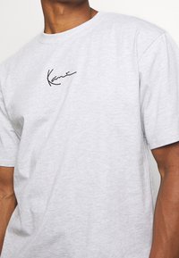Camiseta de algodón gris con mangas cortas, que presenta un logo en cursiva negro en el pecho. Textura suave y ajuste casual.