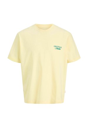 Nastolatek chłopiec uśmiecha się, nosi żółtą koszulkę z napisem, czarne szorty w cienkie prążki, kremowe skarpetki z logo oraz białe trampki, stoi na tle jednolitego tła.