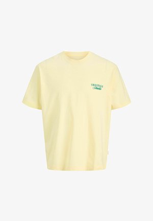 Έφηβος αγόρι χαμογελά, φοράει κίτρινη μπλούζα με κείμενο, μαύρο σορτς με ψιλές ρίγες, κρεμ κάλτσες με λογότυπα και λευκά αθλητικά, στέκεται μπροστά σε απλό φόντο.