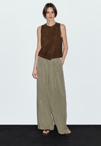 Vrouw die tegen een witte muur staat, gekleed in een mouwloos bruin asymmetrisch topje, olijfgroene wijde broek en zwarte sandalen. Handen in de zakken.