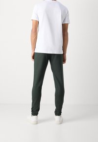 Person in dunkelgrünen Jogginghosen, weißen Sneakers und einem schlichten weißen T-Shirt, der vor einer hellgrauen Wand steht.