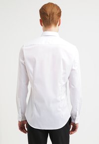 Camicia bianca a maniche lunghe con un design aderente, texture liscia, orlo curvo e polsini con bottone. Nessun motivo o logo visibile.
