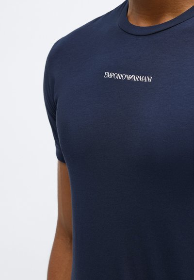 Tumši zilo pieguļošo T-kreklu ar baltu Emporio Armani logotipu centrā uz krūšu puses nēsā persona ar brūnu ādas toni.