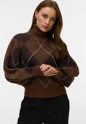 Femme aux longs cheveux bruns portant un pull à col roulé marron et violet foncé à motif argyle et un pantalon noir, mains jointes devant.