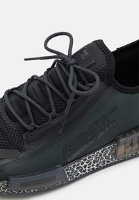 Svart nät-sneaker med texturerade accenter. Har platta snören, gummisula med genomskinlig sektion och upphöjd logotyp på sidan.