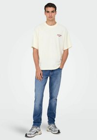 Kratki rukavi krem T-shirt s crvenim logotipom, uparen s plavim trapericama. Model stoji s jednom rukom u džepu, nosi sive tenisice.