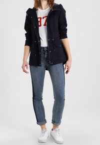 Granatowa kurtka z kapturem i sznurkami, z dwiema przednimi kieszeniami, w połączeniu z białą koszulką z czerwonymi numerami oraz niebieskimi dżinsami z podwiniętymi mankietami.