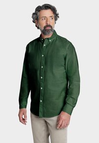 Camisa de lino verde con cuello abotonado y mangas largas, que presenta una textura suave y botones blancos. Combinada con pantalones de color claro.
