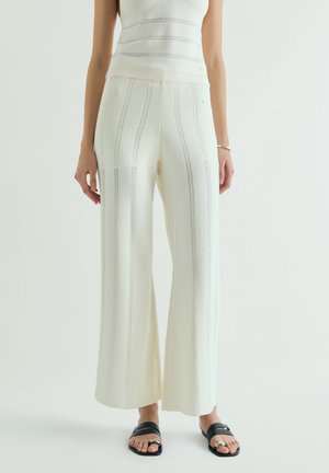 Mujer con pantalones anchos crema de rayas verticales y sandalias negras de punta abierta, de pie sobre un fondo claro y liso.