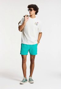 Biele bavlnené tričko s farebnou grafikou na hrudi, doplnené tyrkysovými šortkami. Má na sebe tmavé slnečné okuliare a zelené tenisky.