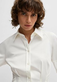 Camicetta bianca con colletto a punta, maniche a raglan corte e una texture liscia. Il tessuto sembra leggero con una vestibilità su misura.
