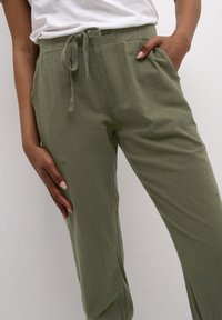 Pantaloni leggeri verde oliva con vita elasticizzata, coulisse regolabile e tasche laterali. Il tessuto ha una finitura morbida e leggermente strutturata.