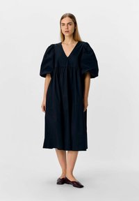 Robe en coton noire avec un décolleté en V, des manches bouffantes et une taille froncée, s'arrêtant juste en dessous du genou. Associée à des chaussures plates marron.