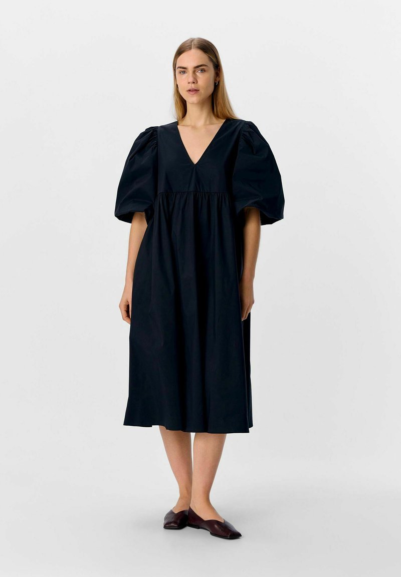 Robe en coton noire avec un décolleté en V, des manches bouffantes et une taille froncée, s'arrêtant juste en dessous du genou. Associée à des chaussures plates marron.