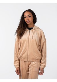 Sudadera con cremallera de terciopelo beige de corte oversize, con capucha ajustable y puños acanalados. Presenta un discreto bordado del logo en el pecho.