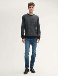 Pull en maille bleu marine foncé avec col rond, manches longues et poignets côtelés, associé à un jean bleu ajusté et des chaussures noires.