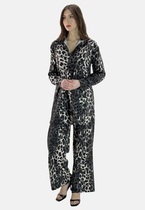Woman wearing loose, leopard-print satin shirt and wide-leg pants with black open-toe heels, standing with hands clasped, looking sideways.