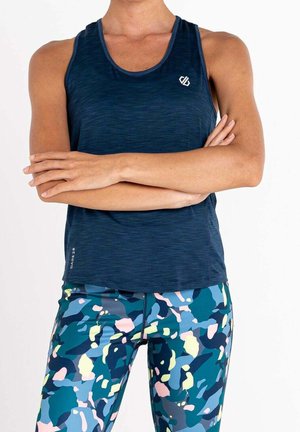 Person trägt ein ärmelloses, marineblaues Sportoberteil und mehrfarbige Leggings mit Camouflage-Muster, Arme vor der Brust verschränkt.