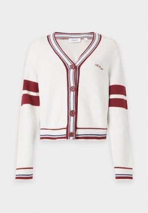 Femme portant des lunettes de soleil foncées, un cardigan blanc avec des rayures rouges et des boutons, un pantalon noir slim et des chaussures argentées à bouts pointus, debout devant un fond uni.