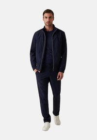 Giacca zip-up blu navy con rifiniture bianche, t-shirt scura, pantaloni abbinati e sneakers bianche. Il tessuto appare liscio con un design aderente.