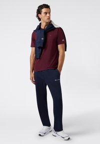 T-shirt bordeaux con un piccolo logo, pantaloni della tuta blu navy e una felpa blu navy e grigia appoggiata sulle spalle. Scarpe sportive bianche.