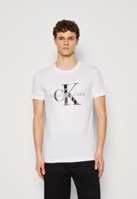 Balts kokvilnas T-krekls ar melnu grafisko logo "CK" un tekstu "Calvin Klein Jeans" zem tā. Īsas piedurknes un apaļa kakla izgriezums.