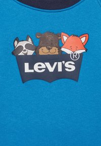 Logotipo de Levi's con un mapache, un oso y un zorro de dibujos animados asomándose por detrás sobre un fondo de tela azul brillante.