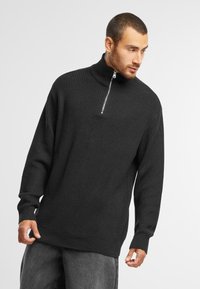Pull en maille noir avec un col haut, fermeture éclair mi-longue, texture côtelée, et manches longues, associé à un pantalon gris ample.