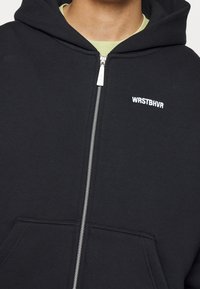 Gros plan d'une personne portant un sweat à capuche noir zippé avec une fermeture éclair argentée et un texte blanc "WRSTBHVR" sur la partie gauche de la poitrine.