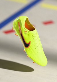 Botín de fútbol de color amarillo neón con una superficie texturizada, swoosh negro con borde rojo y interior naranja. Suela con tacos para mayor agarre.