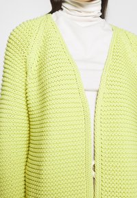 Dziergany kardigan w jasnym żółtym kolorze, z teksturowanym, grubym wzorem. Otwarty z przodu, bez zapięć, noszony na białym golfie.