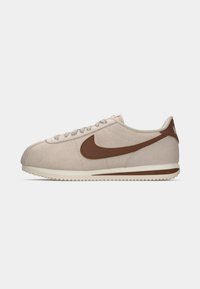 Svetlo bež Nike tenisače s rjavim logotipom swoosh, bež vezalkami in belim podplatom z rjavim lisom, prikazane v stranski profilu na beli podlagi.