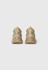 Beige sneakers med skinnende gullremmer og teksturerte såler vist bakfra mot en enkel hvit bakgrunn.
