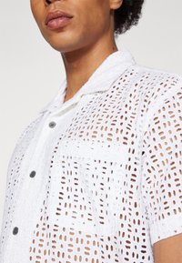 White short-sleeve shirt featuring a textured grid pattern, two buttons, and a chest pocket. Made from lightweight, breathable fabric.