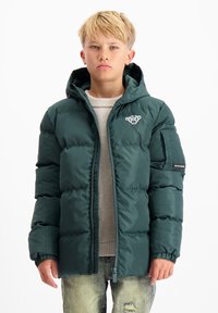 Groene gewatteerde jas met ritssluiting en capuchon, voorzien van een logo op de borst en een verstelbare zoom, gedragen over een grijze trui.