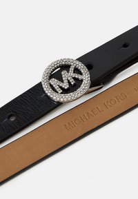 Melnas ādas josta ar teksturētu apdari, kurai ir apaļa sprādze, kas rotāta ar kristāliem un ar MK logo. Iekšpusē ir izsistīts "Michael Kors" zīmols.