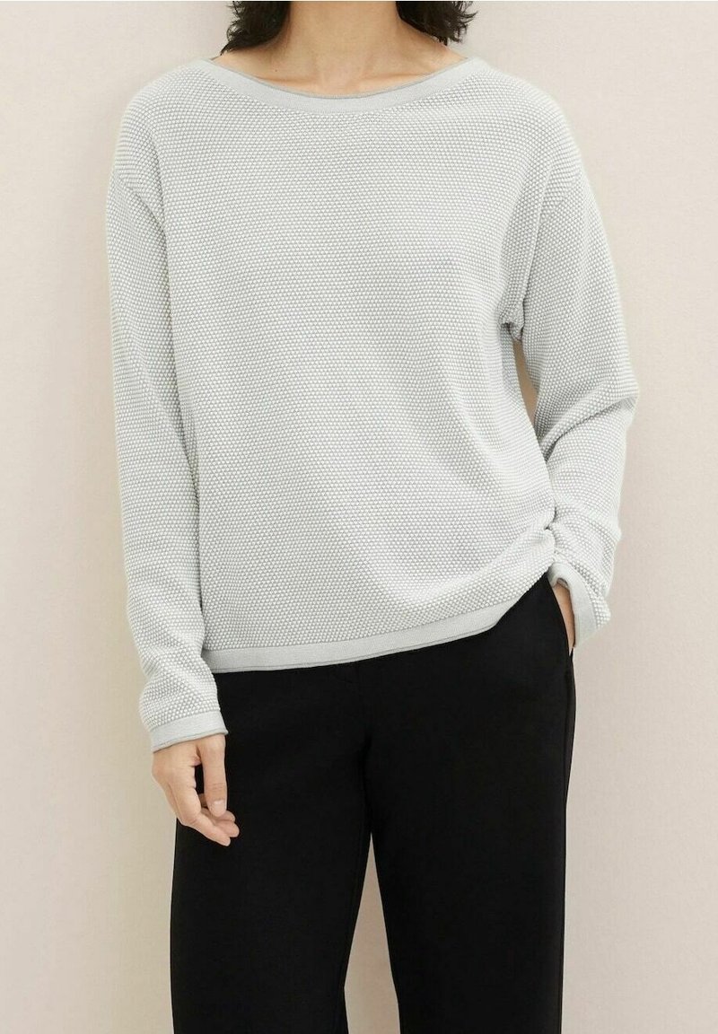 Personne portant un haut à manches longues gris clair texturé et un pantalon noir, debout avec une main dans la poche, sur un fond neutre.