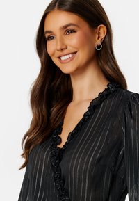 Blusa nera con strisce verticali argentate, scollo arruffato e tessuto trasparente. Presenta maniche lunghe a sbuffo e un leggero lucido.