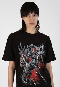 Tricou negru din bumbac, prezentând un grafic complex cu un cal legat, pe un fundal roșu și negru, cu litere albe îndrăznețe deasupra.